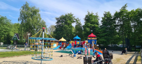 Niños juegan en un colorido parque infantil en Vakantiepark 't Urkerbos, rodeados de árboles y familias.