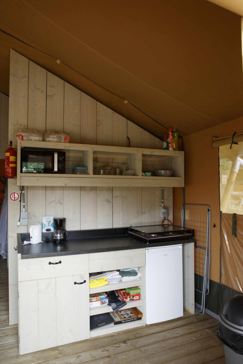 Kitchen area inside a Safari tent lodge at Costa Kabrita in the Netherlands, with shelves, stove, and mini fridge.