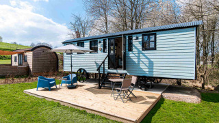 Shepherd Lodge glamping con carrozza blu, terrazza in legno, sedie, ombrellone e prato verde intorno.