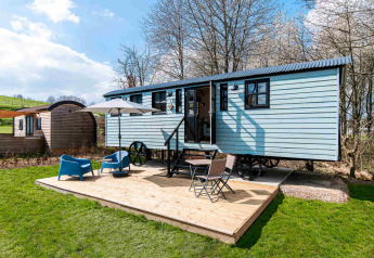 Shepherd Lodge glamping met blauwe woonwagen, houten terras, stoelen, parasol en groene tuin.