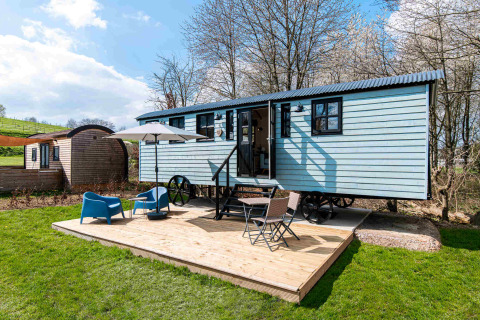 Shepherd Lodge glamping met blauwe woonwagen, houten terras, stoelen, parasol en groene tuin.