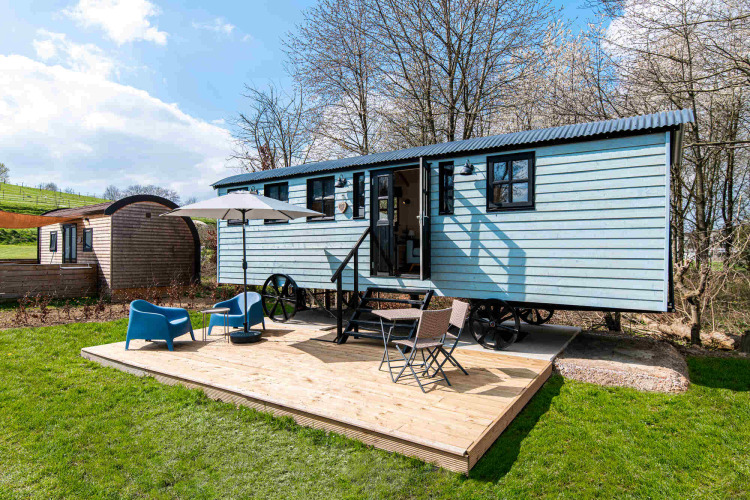 Glamping Shepherd Lodge con cabaña azul, terraza de madera, sillas, sombrilla y césped alrededor.