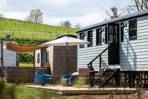 Glamping-Bereich bei Shepperd Lodge mit Holzhütten, Sitzgelegenheiten und Sonnenschirm auf einer Wiese.