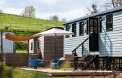 Espace glamping extérieur à Shepperd Lodge avec cabanes en bois, mobilier de jardin et parasol sur pelouse.