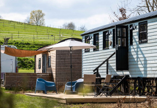 Glamping-Bereich bei Shepperd Lodge mit Holzhütten, Sitzgelegenheiten und Sonnenschirm auf einer Wiese.