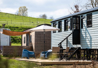 Outdoor glamping area at Shepperd Lodge featuring wooden cabins, garden furniture, and a parasol on grass.