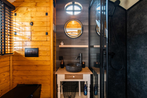 Baño moderno en Shepperd Lodge glamping con paredes de madera, espejo redondo y lavabo de piedra negra.