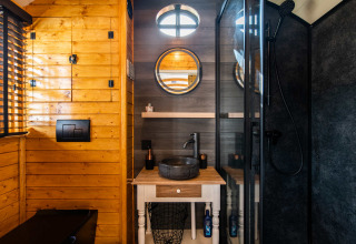 Baño moderno en Shepperd Lodge glamping con paredes de madera, espejo redondo y lavabo de piedra negra.