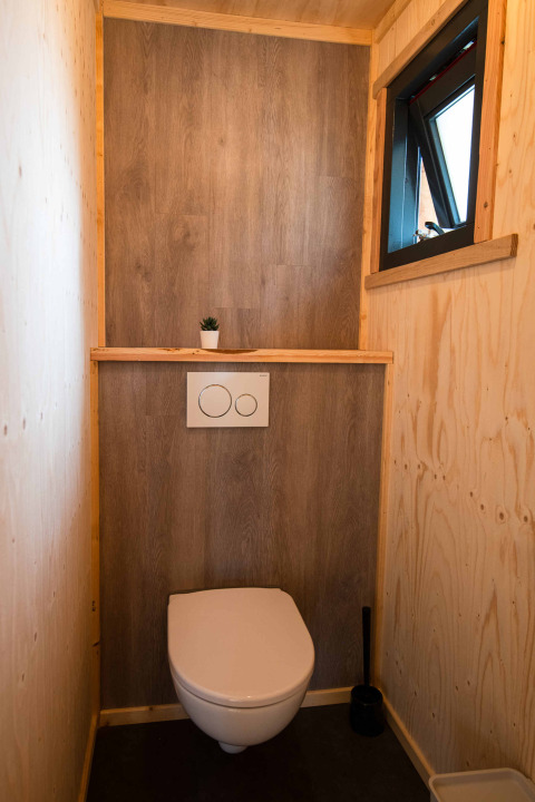 Baño moderno y compacto con paredes de madera, pequeña ventana y planta en la tiny house Jachthut en Camping Vinkenhof.