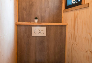 Baño moderno y compacto con paredes de madera, pequeña ventana y planta en la tiny house Jachthut en Camping Vinkenhof.