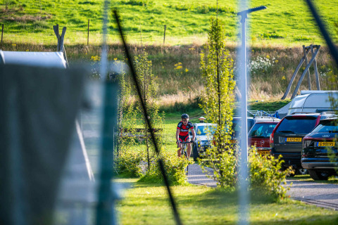 Un ciclista arriva al Camping Vinkenhof a Limburg, Paesi Bassi, tra auto parcheggiate e paesaggio verde.