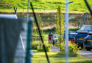 A cyclist arrives at Camping Vinkenhof holiday park in Limburg, Netherlands, among parked cars and greenery.