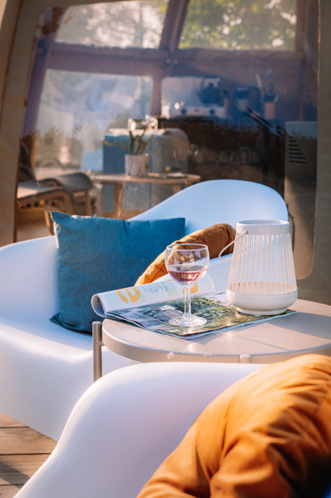 Zona de descanso en Panorama Dome, Camping Vinkenhof, con copa de vino, revista y cojín azul brillante.