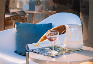 Zona de descanso en Panorama Dome, Camping Vinkenhof, con copa de vino, revista y cojín azul brillante.