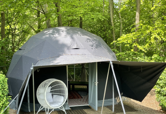Geodetische Dome Tent in het bos, met een witte hangstoel vooraan en omgeven door groene bomen.