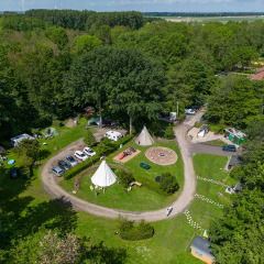 Vista aérea de un alojamiento glamping rodeado de bosque, con tiendas, coches, caminos y áreas recreativas.