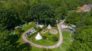 Vista aérea de un alojamiento glamping rodeado de bosque, con tiendas, coches, caminos y áreas recreativas.