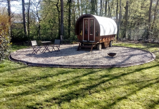 Una Gipsy Wagon tiny house en el bosque, con una pequeña mesa y dos sillas sobre una zona de grava.