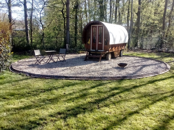 Een Gipsy Wagon tiny house in het bos, met een klein tafeltje en twee stoelen op een grindterrein.