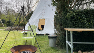 Escena al aire libre con tienda tipi, fogata, dos sillas verdes, mesa y leña sobre césped.