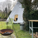 Udendørs scene med en tipi-telt, bålplads, to grønne stole, bord og brænde på græsplæne.