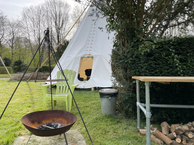 Scène extérieure avec une tente tipi, un foyer, deux chaises vertes, une table et du bois.