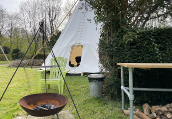 Escena al aire libre con tienda tipi, fogata, dos sillas verdes, mesa y leña sobre césped.