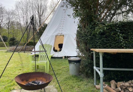 Escena al aire libre con tienda tipi, fogata, dos sillas verdes, mesa y leña sobre césped.