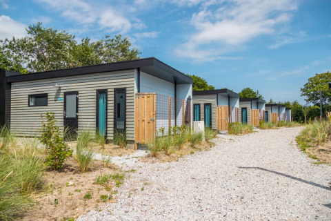 Rij moderne glampinghuisjes in een vakantiepark met grindpad en duinplanten, onder een blauwe lucht.