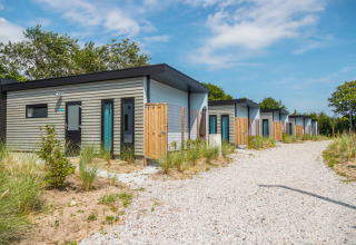 Row of modern glamping cabins in a holiday park, with gravel path and sandy landscaping, under blue sky.