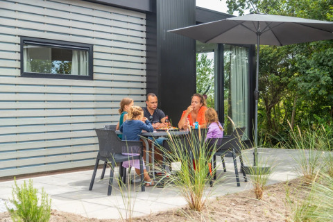 A family enjoys an outdoor meal together at a glamping holiday park, sitting under a large patio umbrella.