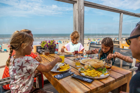 Familia disfruta de una comida en una terraza de restaurante junto a la playa con vista al mar y cielo azul.