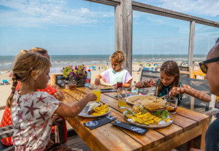 Family enjoys lunch at a wooden table in a beachside restaurant, with ocean view and blue sky.