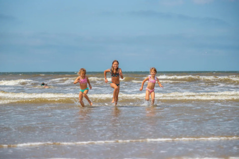 Tres niños corren alegres por la orilla del mar en un parque vacacional con opciones de glamping.