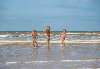 Tre børn løber glade i havet ved en strand under en blå himmel, taget fra et feriepark med glamping.
