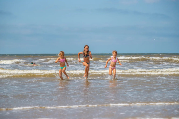 Drie kinderen rennen lachend door de branding bij een vakantiepark met glamping accommodaties.