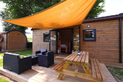 Glamping-Unterkunft aus Holz mit Terrasse, Gartenmöbeln und orangefarbener Sonnensegelplane.
