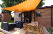 Glamping-Unterkunft aus Holz mit Terrasse, Gartenmöbeln und orangefarbener Sonnensegelplane.