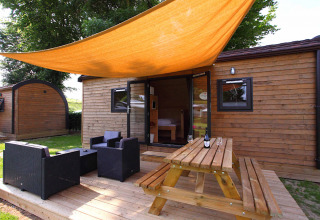 Glamping-Unterkunft aus Holz mit Terrasse, Gartenmöbeln und orangefarbener Sonnensegelplane.