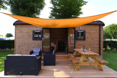 Casa glamping con terraza de madera, zona lounge, mesa de picnic y toldo naranja bajo árboles verdes.