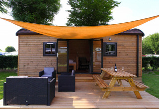 Casa glamping con terraza de madera, zona lounge, mesa de picnic y toldo naranja bajo árboles verdes.