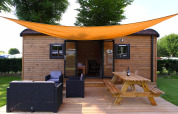 Casa glamping con terraza de madera, zona lounge, mesa de picnic y toldo naranja bajo árboles verdes.