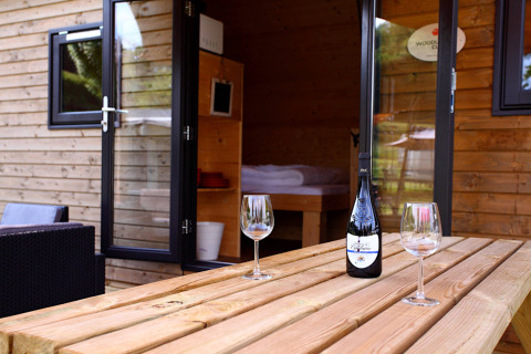 Cabaña de glamping con puertas abiertas, botella de vino y dos copas en mesa de madera exterior.