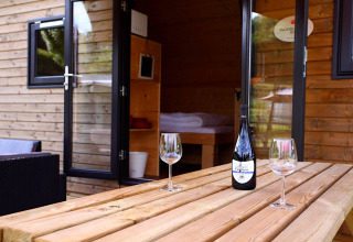 Træhytte glamping med åbne døre, vinflaske og to vinglas på terrassebord, hyggelig stemning.