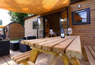 Outdoor glamping cabin with picnic table, wine bottle, and glasses under a sunshade canopy.