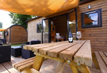 Sistemazione glamping all’aperto con cabina in legno, tavolo da picnic, vino e bicchieri all’ombra.
