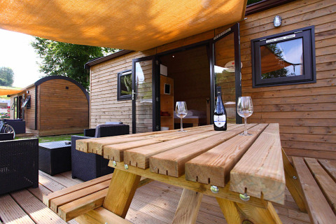 Udendørs glampingområde med træhytte, picnicbord, vinflaske og vinglas under en solsejl.