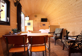 Acogedora cabaña de glamping con paredes de madera, mesa de comedor, TV, cama doble y sillones estampados.