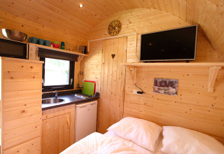 Interno accogliente di una cabina glamping con pareti in legno, angolo cottura, letto e TV a parete.
