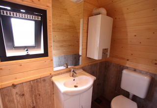 Baño con paredes y suelo de madera, lavabo, inodoro, espejo, calentador de agua y ventana en glamping.